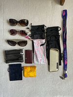 Top-down view of three ladies' sunglasses, multiple black sunglasses pouches, zippered pouches, small fabric bags, and a patterned fabric case with a purple lanyard.