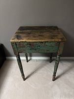 Front view of vintage side table showing heavily distressed green and black paint and natural wood tabletop with visible split