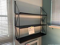Front view of the 3 tier black metal and wood bookshelf mounted on a pale blue wall next to a window with blinds.