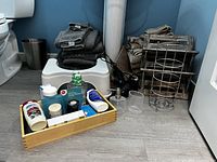 Wide view showing towels, white plastic step stool, metal toilet paper stand, metal shelf, blue tray with toiletries including lotions, razors, and other personal care items on bathroom floor