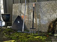 Garden tools including a leaf rake, dethatching rake, rotary cultivator, and metal rake handle visible, against a stone wall background