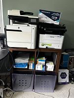 Full view of the two printers on the wooden shelf unit, showing the extra toner cartridge box and paper supplies, plus storage bins below the shelf.