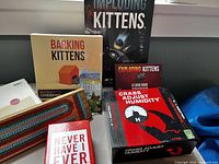 Photo showing multiple card and board game boxes including Barking Kittens, Imploding Kittens, Exploding Kittens, Streaking Kittens, Crabs Adjust Humidity, and Never Have I Ever placed on a flat surface.