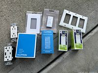Collection of electrical items including Lutron Caséta Wireless Dimmer kit, Leviton 3-way switches, electrical outlets, and switch plates laid out on concrete surface.