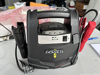 Front view of black Schumacher Instant Power battery jump starter showing handle, LED light panel, switch, USB port, and red and black alligator clip cables attached.