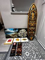 Full set of seven items including tiki surfboard, two tiki masks, and four framed paintings arranged against a wall.