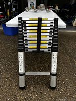 Folded telescopic aluminum ladder showing multiple rungs stacked with a black strap holding them together.