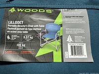 Label showing brand WOODS, model Lillooet portable director's chair with table, specs include 300 lb capacity, 16.5 inch seat height, lightweight aluminum frame, made in Canada.