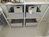 Front view of two gray and white shelving units side by side, showing metal frames and gray shelving boards. Storage boxes present but not included.