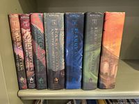Original 7 Harry Potter hardback books shown upright with spines visible, colors and titles clear including Philosopher's Stone, Chamber of Secrets, Prisoner of Azkaban, Goblet of Fire, Order of the Phoenix, Half Blood Prince, Deathly Hallows.
