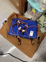 Folded brown heavy duty canvas tarp with orange bungee cords and blue tarp on top, hooks visible