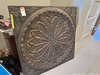 Full view of the large square metal wall art with detailed embossed pattern leaning against furniture.