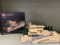Front view of the fully assembled Lego Architecture Fallingwater set with box included, showing the multi-level tan brick house, surrounding greenery, and flowing water feature.