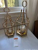 Pair of matching gold metal candle holders with white candles in glass holders, on white surface near window.
