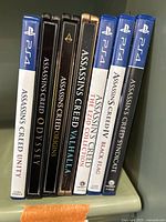 Seven PS4 Assassin's Creed game cases standing upright, showing titles: Unity, Odyssey, Origins, Valhalla, The Ezio Collection, Black Flag, Syndicate.