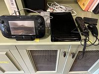 WiiU console with gamepad, two remotes and cables on cabinet top.
