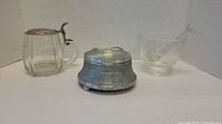 Three items: glass mug with metal lid, glass mortar with pestle, and metal music box, showing overall contents of the lot