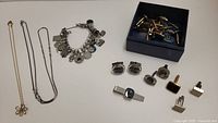 Wide view of cufflinks, tie pins and charm bracelet in a box and laid out on a surface.