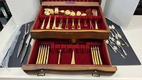 Top view of the wooden case showing the two drawers with gold colored flatware including knives, forks, spoons, and serving pieces with red velvet lining.