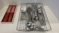 Photo showing 2 steak knives with wooden handles in original box, beside wire tray with forks, knives, spoons arranged