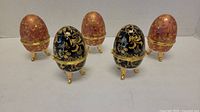 Five porcelain egg containers displayed, two black with gold and blue decoration, three reddish-brown with gold designs, all standing on gold-colored legs.