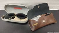 Two Porsche Carrera sunglasses sets with cases; one white hard shell case holding sunglasses and extra lenses, other set with brown leather case
