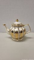 Front view of teapot showing gold and ivory alternating vertical stripes with floral decorations and gold handle and spout tip.
