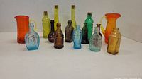 Overall view of 14 colorful glass bottles including blue, green, amber, and brown colors, some with cork stoppers and various shapes and textures, plus two orange pitchers at the back