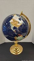 Front side of the globe showing North America with countries made of different colored stones and shells on a deep blue ocean background, mounted on a gold-tone metal stand.