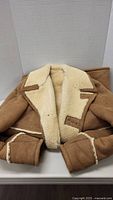 Medium length brown suede shearling coat laying on table showing cream shearling lining on collar and inside.
