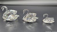 Three clear crystal swan figurines of different sizes placed on a reflective surface, showing their faceted cuts and details.