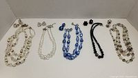 Five vintage necklace sets displayed in a white box, with matching earrings for each set. Variety of beads including faux pearls, blue glass, black beads, and multicolor glass.