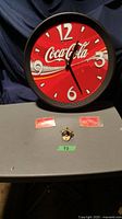 Photo showing Coca-Cola wall clock, metal bottle opener and two pack of multi-piece fridge magnets arranged on a gray surface against dark background