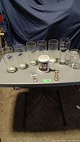Photo showing all items on table including six pilsner glasses, one Labatt Blue pint glass, one coffee mug, a blue light keychain opener combo, and a vintage metal bottle opener. Items arranged in a row with visible branding.