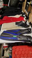 Photo showing two pairs of diving flippers, one black and blue Oceanic brand and the other large white Liberator flippers, alongside wetsuits.