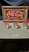 Photo showing three vintage Coca-Cola glasses with red text and a framed vintage Coca-Cola sign on a table.