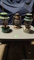 Three lanterns sitting on a table; two green Coleman lanterns and one copper decorative lantern in the middle.