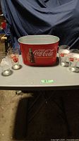 Full lot overview showing red Coca-Cola metal ice tub and five Coca-Cola themed drinking glasses arranged on table