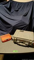 Photo showing the closed orange Plano fishing tackle case alongside the closed light grey Pelican 1450 hard case on a table.