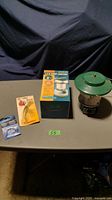 Photo of the camping lot with the LED lantern in box, Coleman propane lantern, Sure Fire fishing lure, and steel leaders set arranged on a table.