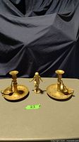 Pair of metal candle holders with attached bells and solid metal figure between them on a table.