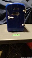 Front view of blue Sunbeam mini fridge with oval window and silver handles on table.