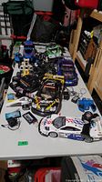 Several remote control cars on a table with multiple wired chargers and remote controllers spread around them.