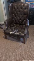 Dark brown faux leather wing back chair with cracked and peeling leather seat, button tufted backrest, wooden frame and legs with nailhead trim.