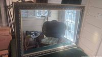 Full view of large rectangular gilded framed mirror reflecting a wicker chair and window.