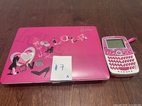 Pink Barbie-themed educational toy laptop alongside a Barbie-themed toy phone on wooden floor.