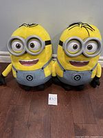 Front view of two Despicable Me Minion Dave plush toys standing side by side on a floor with a price tag in front.