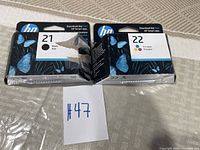 Photo of HP 21 Black and HP 22 Tri-color ink cartridges in sealed packaging with a handwritten note number 47 for lot identification.