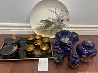 Full view of Japanese tea sets including black lacquer tea cups on trays, cobalt blue decorative bowl, lidded jars, and a large plate with bird and floral designs.