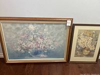 Two framed floral paintings side by side on a wooden floor against a white wall. The larger painting on the left depicts a bouquet in a glass vase and is signed. The smaller painting on the right has a brown vase with white and yellow flowers.